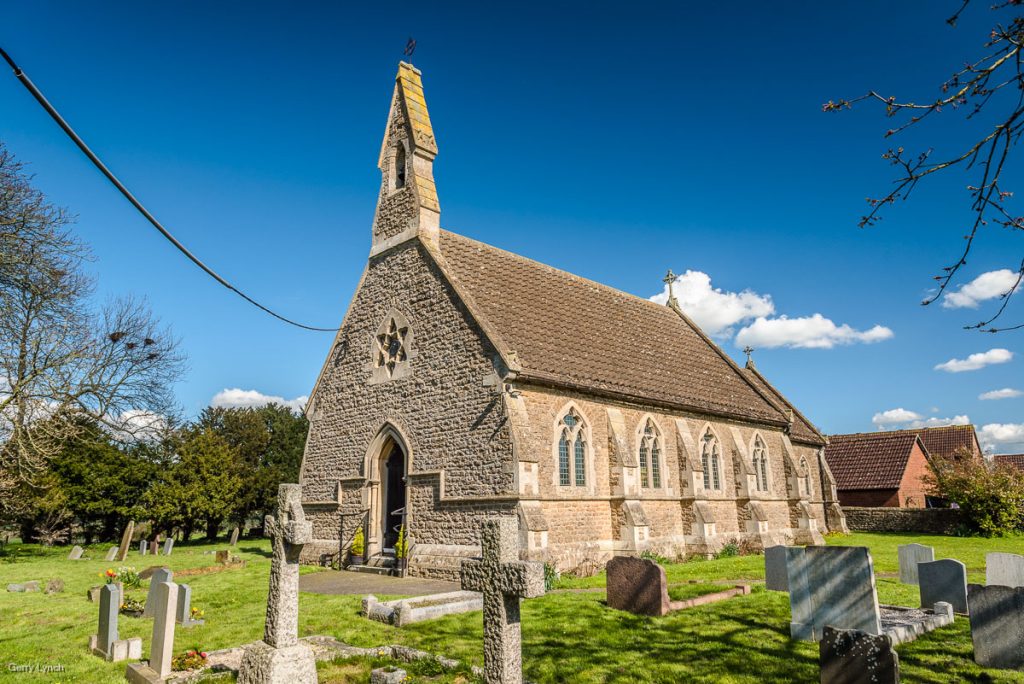 A stone church with a steeply pitched roof and a small pointed tower, featuring arched windows, stands under a bright blue sky with scattered white clouds. The church is surrounded by a well-kept graveyard with various tombstones and crosses, set amidst green grass and trees. A power line stretches across the sky, and a small brick building is visible in the background.