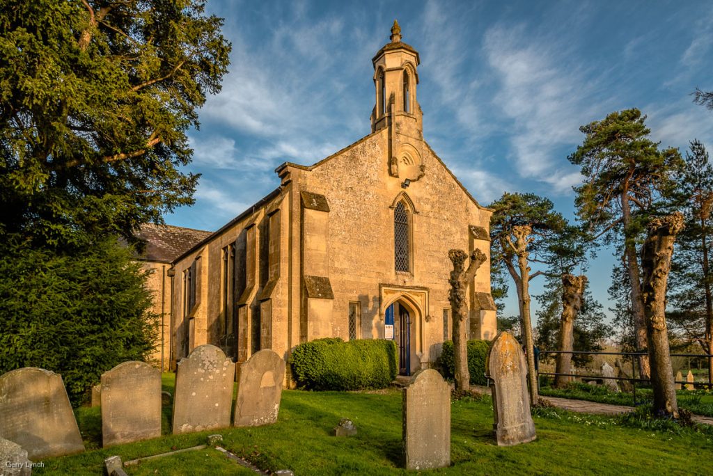A historic stone church with a small tower and arched windows, surrounded by a well-maintained graveyard with weathered tombstones. The scene is framed by lush green trees and bushes, with a clear blue sky and soft clouds in the background, bathed in warm sunlight.