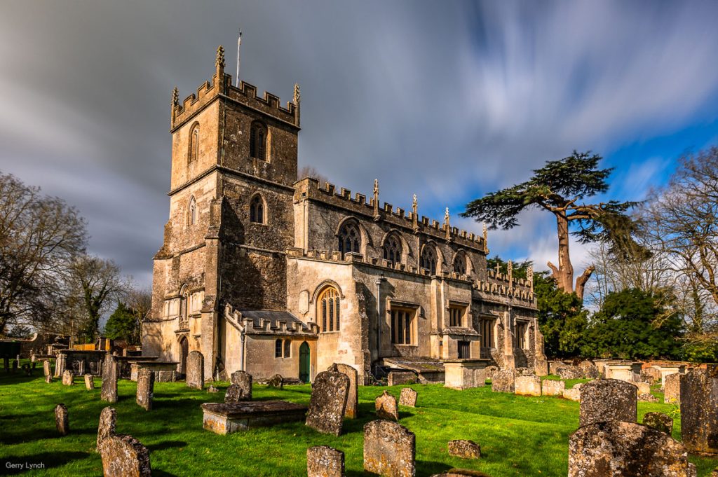 A dramatic view of an ancient stone church with a towering, crenelated tower, surrounded by a historic graveyard filled with weathered tombstones. The church features intricate Gothic windows and is framed by tall, leafless trees and a large evergreen under a sky with wispy clouds and patches of blue. The scene is bathed in soft sunlight, casting gentle shadows across the grass.