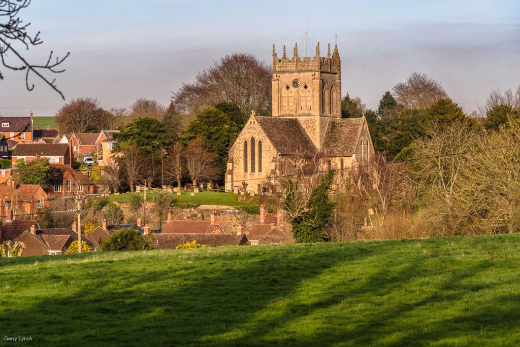  A picturesque view of a historic stone church with a tall, ornate tower, set against a backdrop of lush green trees and a small village with red-brick houses. The scene is framed by a grassy hill in the foreground, with bare tree branches adding a natural touch to the serene landscape.