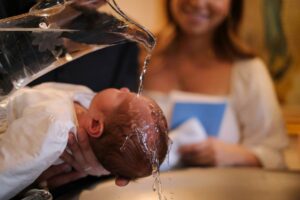Close-up of an infant being baptised: water is poured from a glass ewer over the baby's head, held over a font by an adult's hand. A smiling woman holding a blue book blurs in the background.