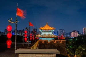 Night photograph of Xi'an's ancient city wall fortifications. A traditional Chinese gate tower with golden glazed tile roof is dramatically lit in the centre, flanked by red paper lanterns and red flags with gold poles mounted on the crenellated stone walls. The twilight sky is deep blue; the modern Xi'an skyline glows in the background with numerous high-rise buildings. A stone parapet runs along the foreground wall. Photograph © Gerry Lynch.